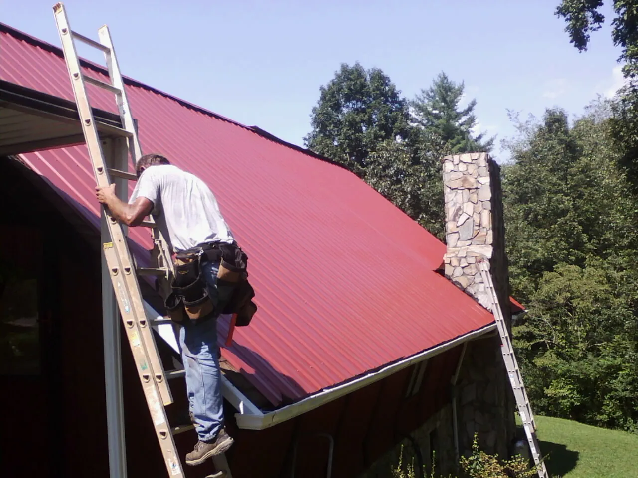 Roofer on a ladder beside a red metal roof and stone chimney — Soffit & Fascia Repair by Roofing Brothers in Fort Mitchell